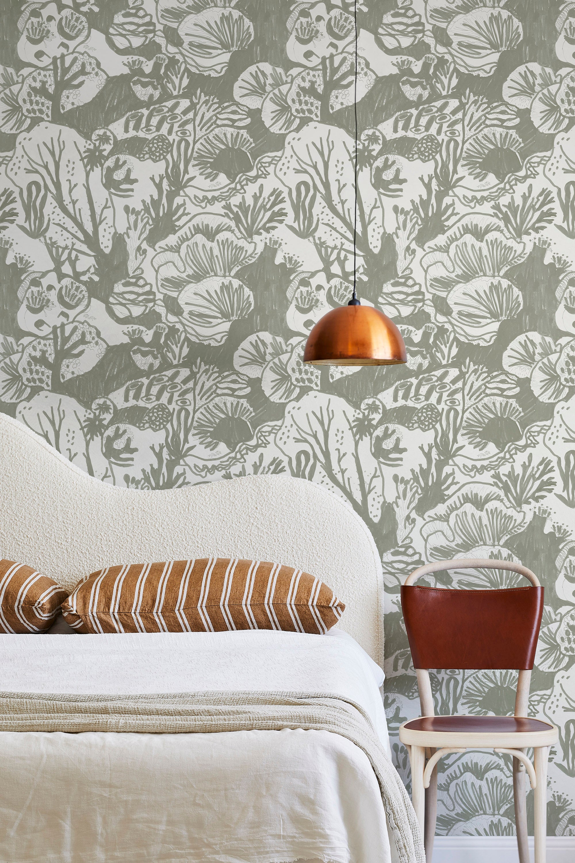 A modernist bed, hanging lamp and chair stand in front of a wall papered in a playful reef print in sage and white.