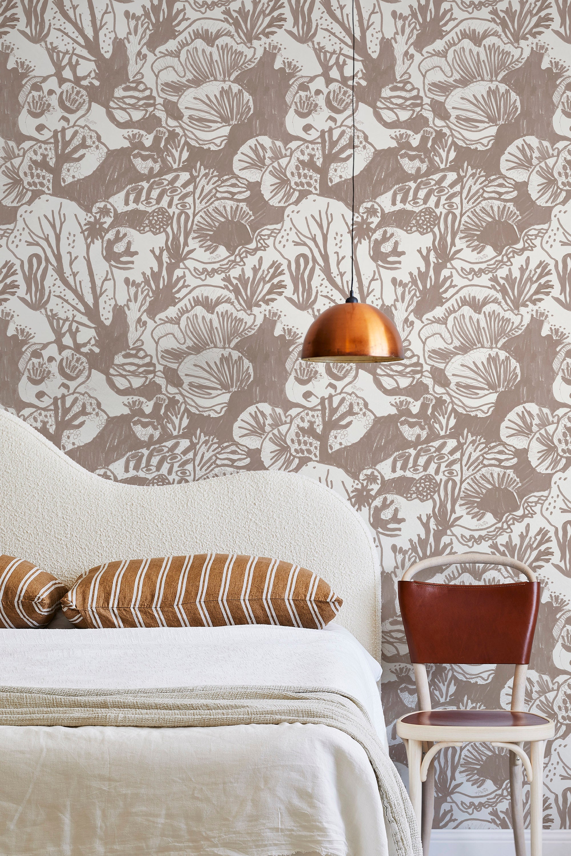 A modernist bed, hanging lamp and chair stand in front of a wall papered in a playful reef print in tan and white.