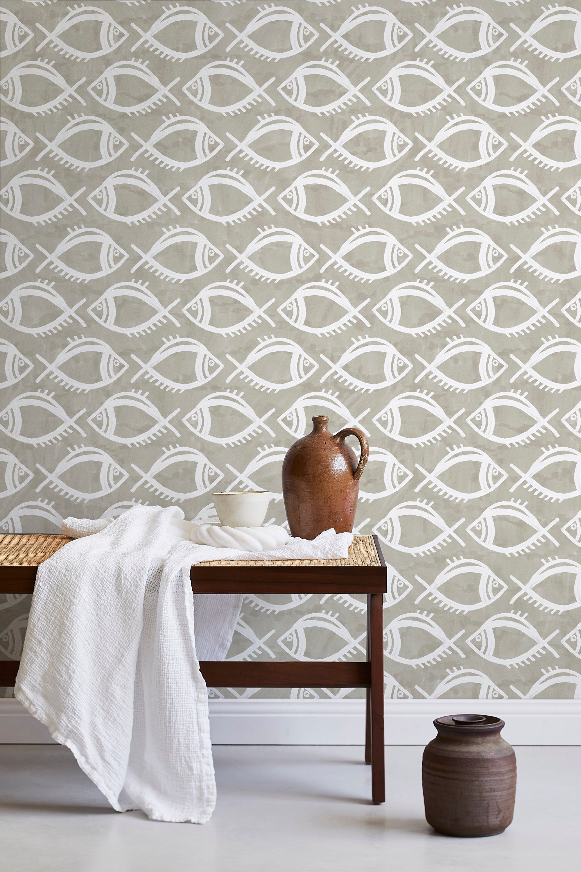 A bench with a towel and clay vases stands in front of a wall papered in a playful fish print in white and greige.