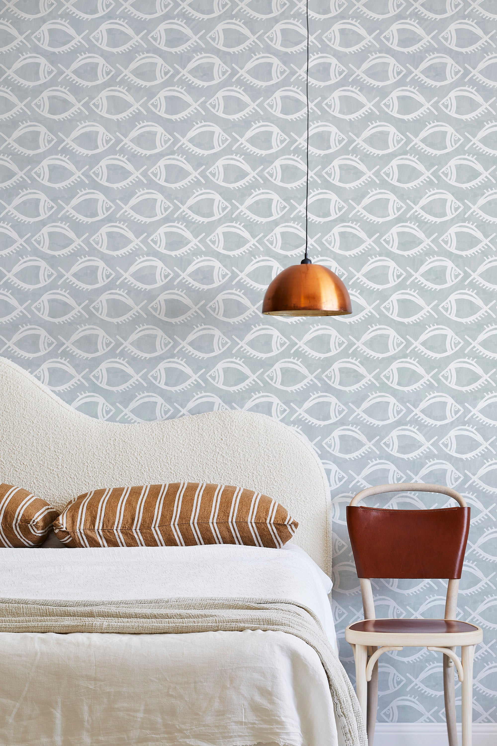 A modernist bed, hanging lamp and chair stand in front of a wall papered in a playful fish print in white and light blue.