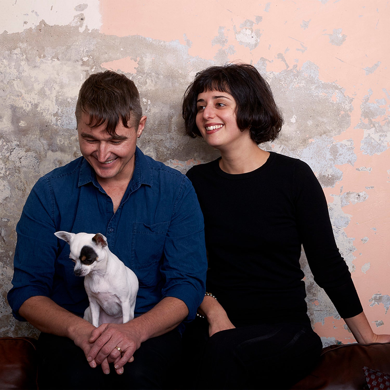 A man and woman both sitting on the ground with a chihuahua, all seated in front of a wall covered in crumbly peach plaster.