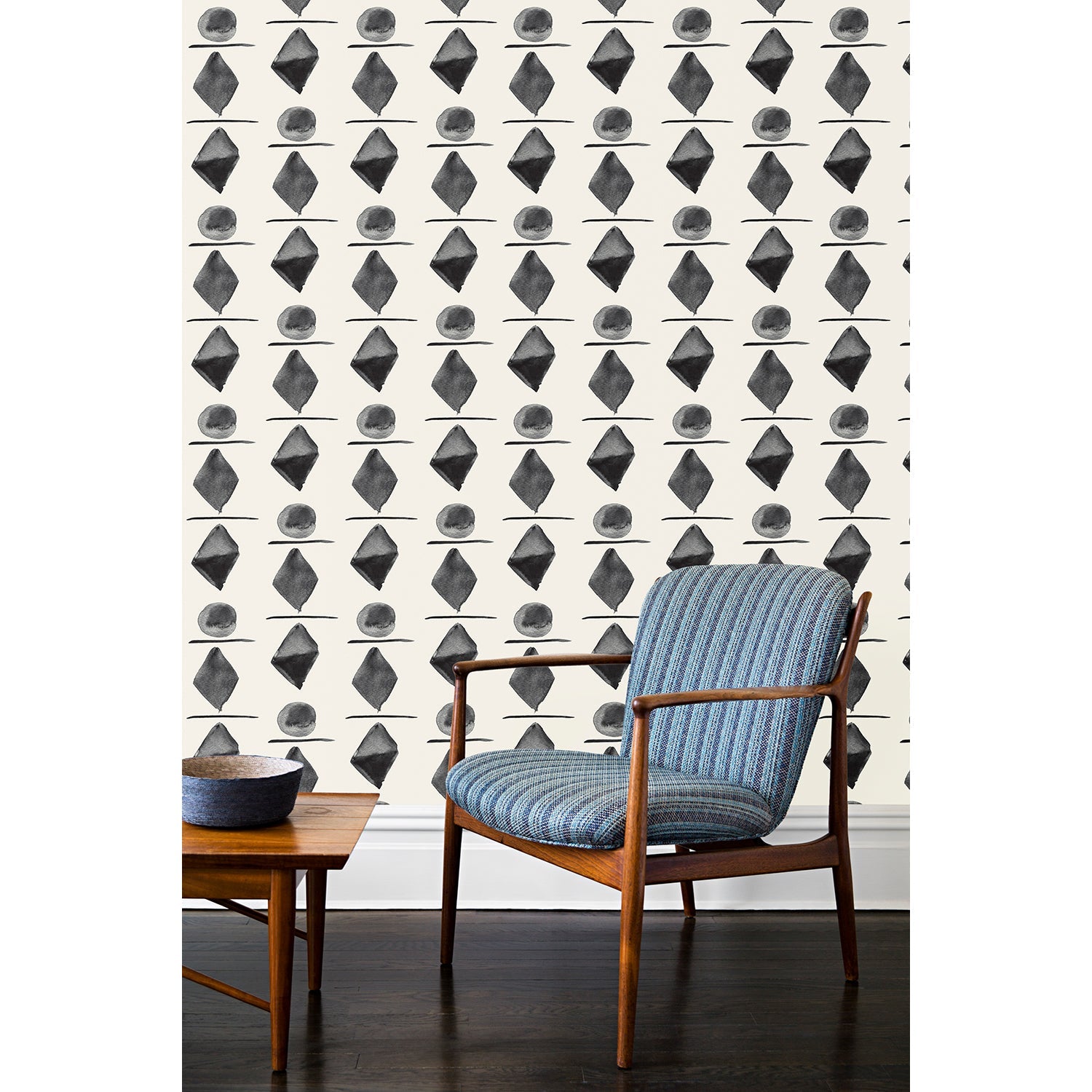 A wooden chair and coffee table in front of a wall papered in a large-scale pattern of black watercolored diamonds, circles and lines on a cream background. 