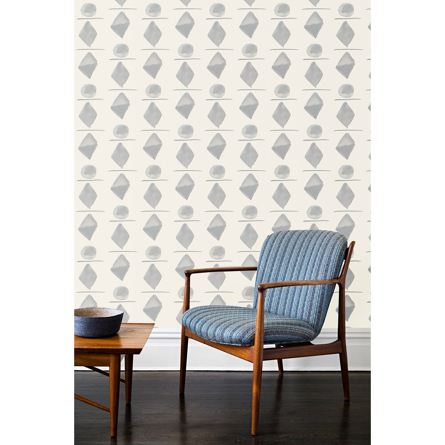 A wooden chair and coffee table in front of a wall papered in a large-scale pattern of gray watercolored diamonds, circles and lines on a cream background. 