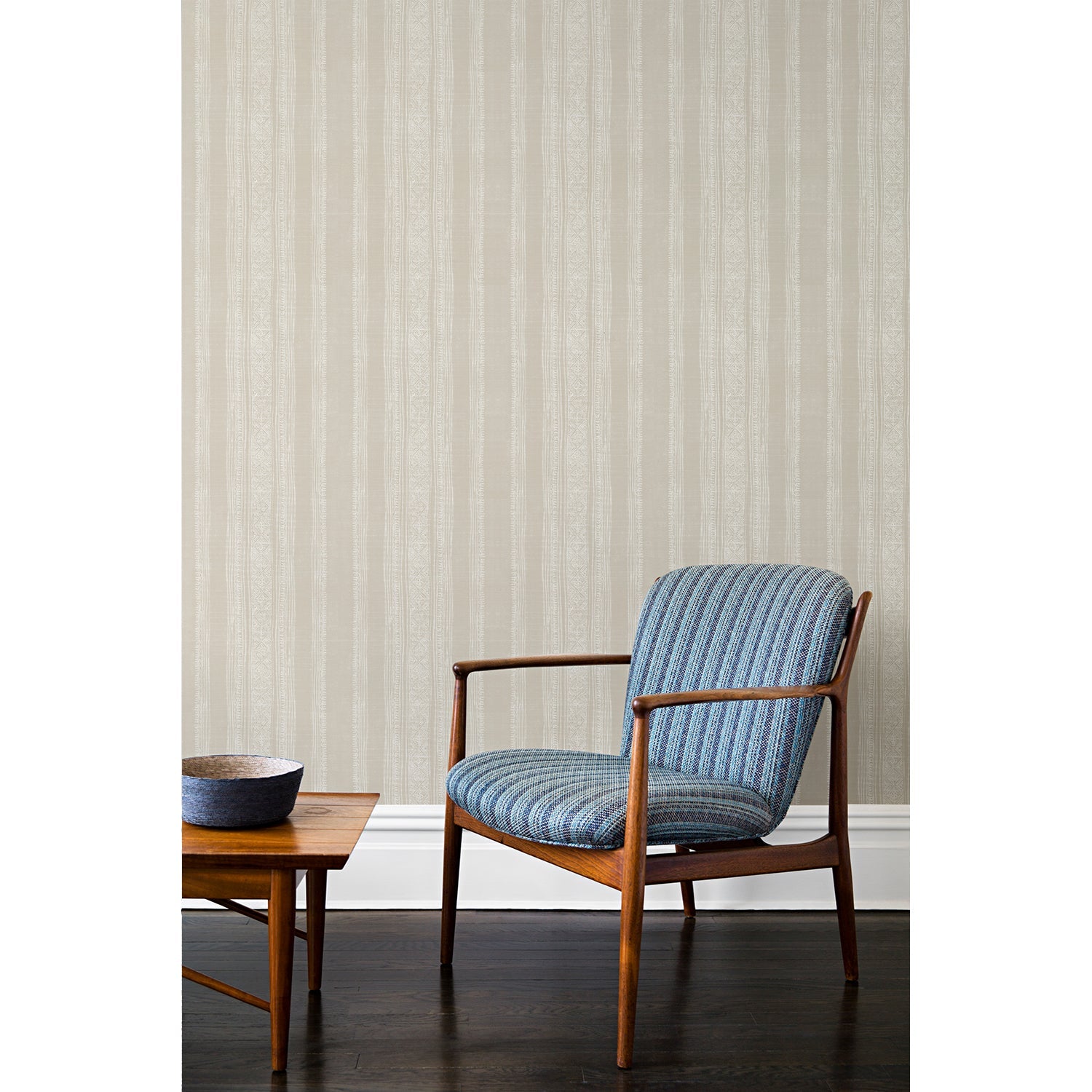 An armchair and coffee table in front of a wall papered in a striped pattern of intricate white lines over a washed beige background.