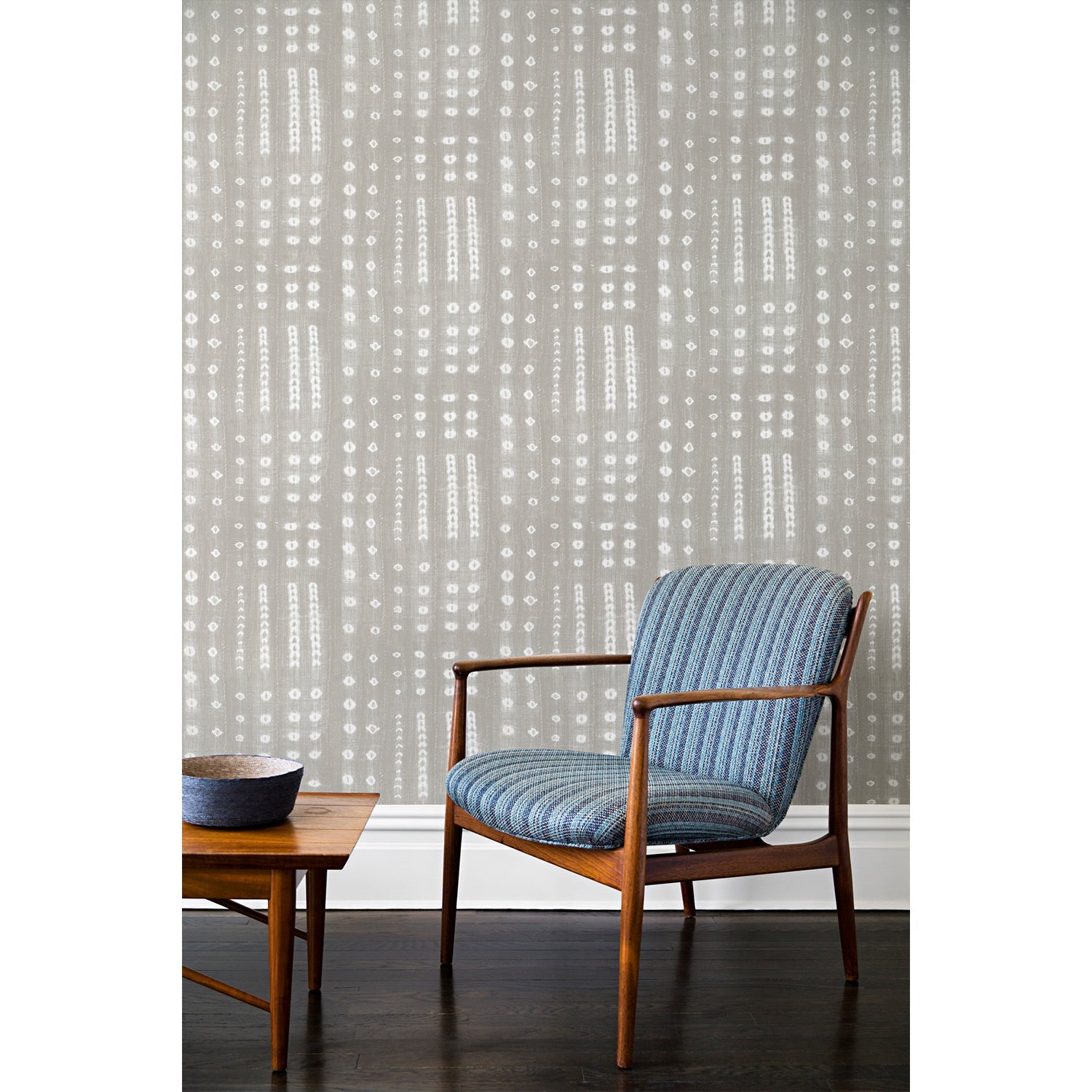 A wooden chair and coffee table in front of a wall papered in a striped print inspired by Japanese shibori on a washed gray background.