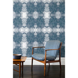 A wooden chair and coffee table in front of a wall papered in a dense hand-drawn symmetrical pattern in shades of blue on a white background.