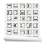 Partially unrolled wallpaper roll with rows of block-printed moons in various phases. Moons are black on a white background.