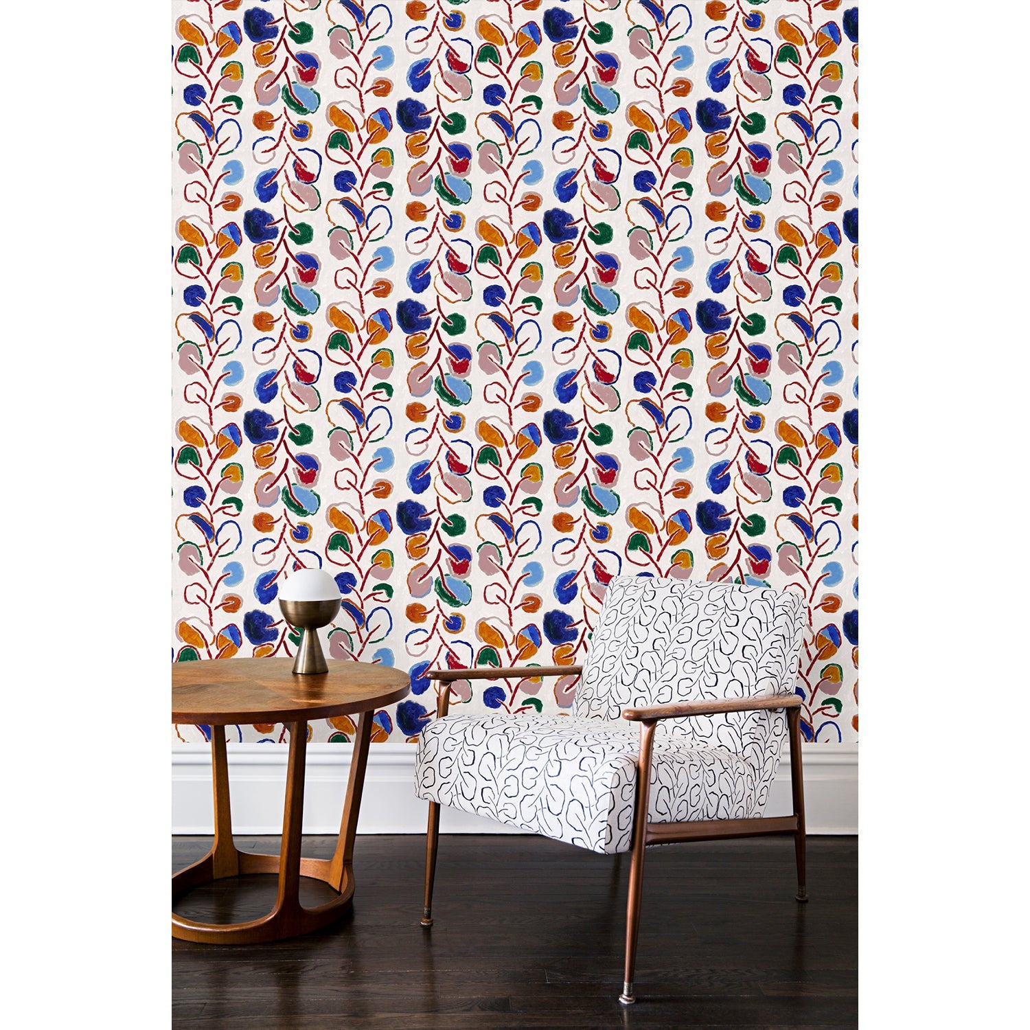 An armchair and coffee table in front of a wall papered in repeating rows of colorful leaf prints in shades of red, purple and blue on a cream background.