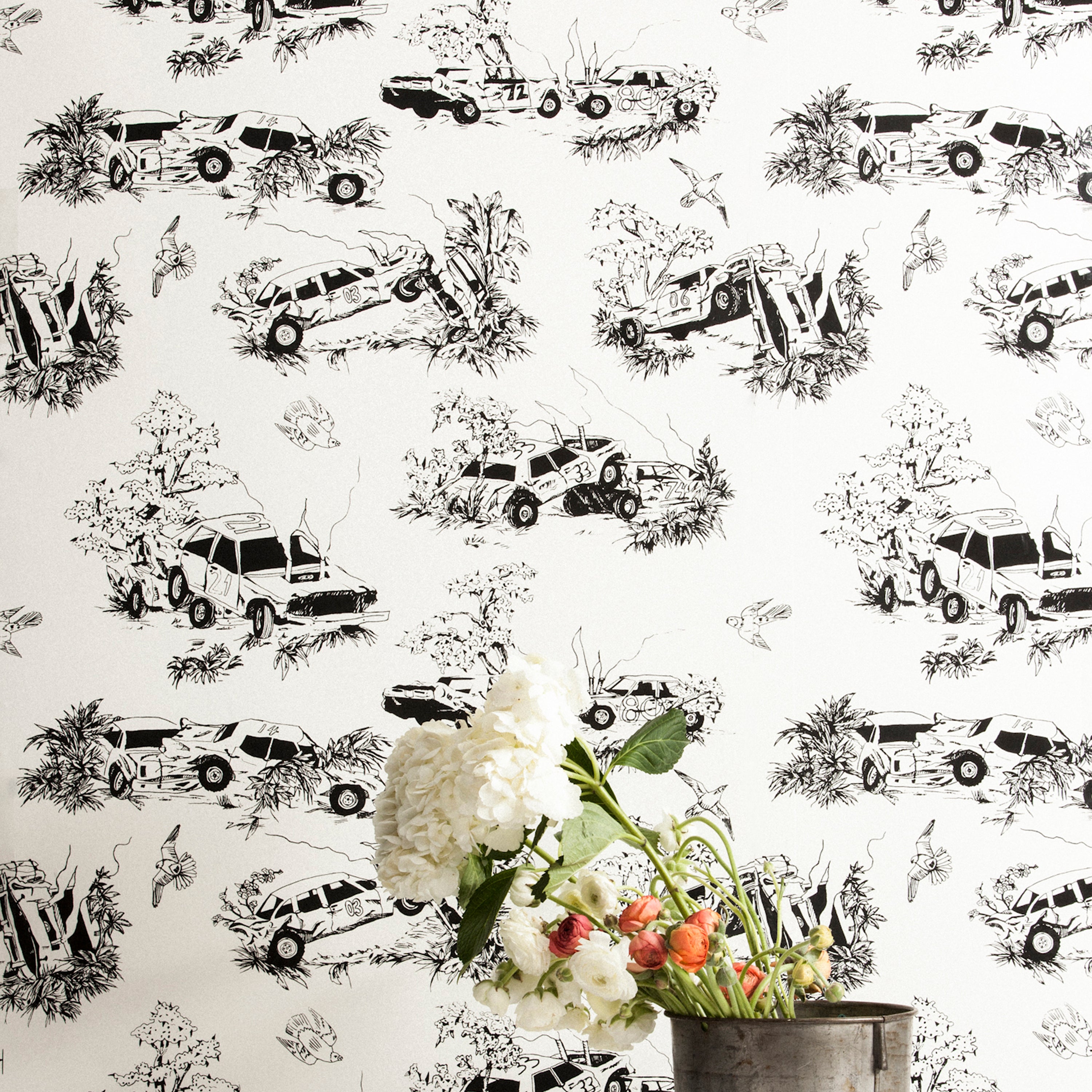 A vase of flowers stands in front of a wall covered in a playful illustrated car crash print in black on a white field.