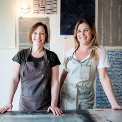 Two smiling women, one older and one younger, wearing aprons and standing with their hands resting on a large drafting table.