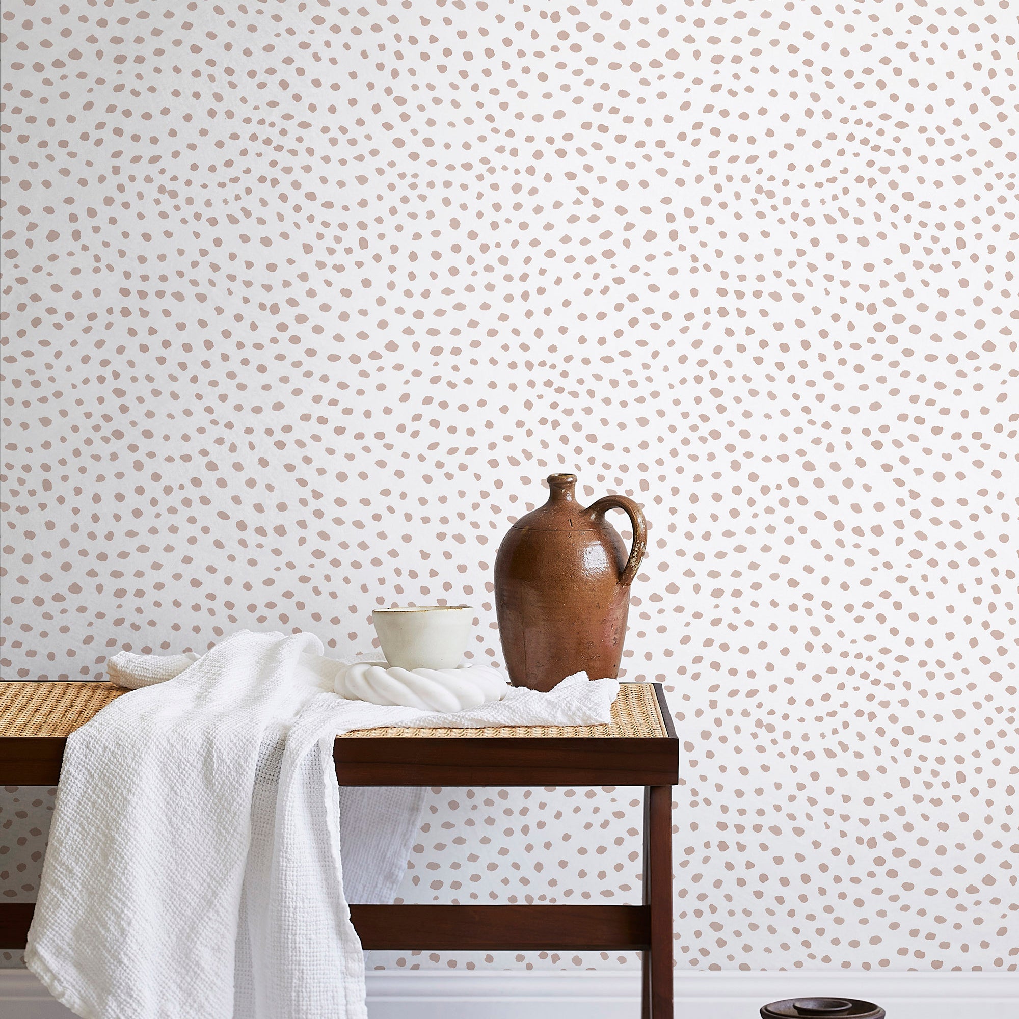 A bench with a towel and clay vases stands in front of a wall papered in a scalloped dot print in blush and white.