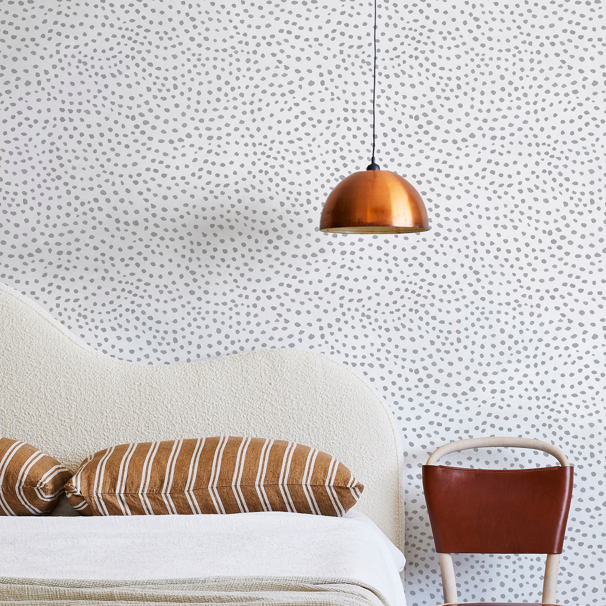 A modernist bed, hanging lamp and chair stand in front of a wall papered in a scalloped dot print in gray and cream.