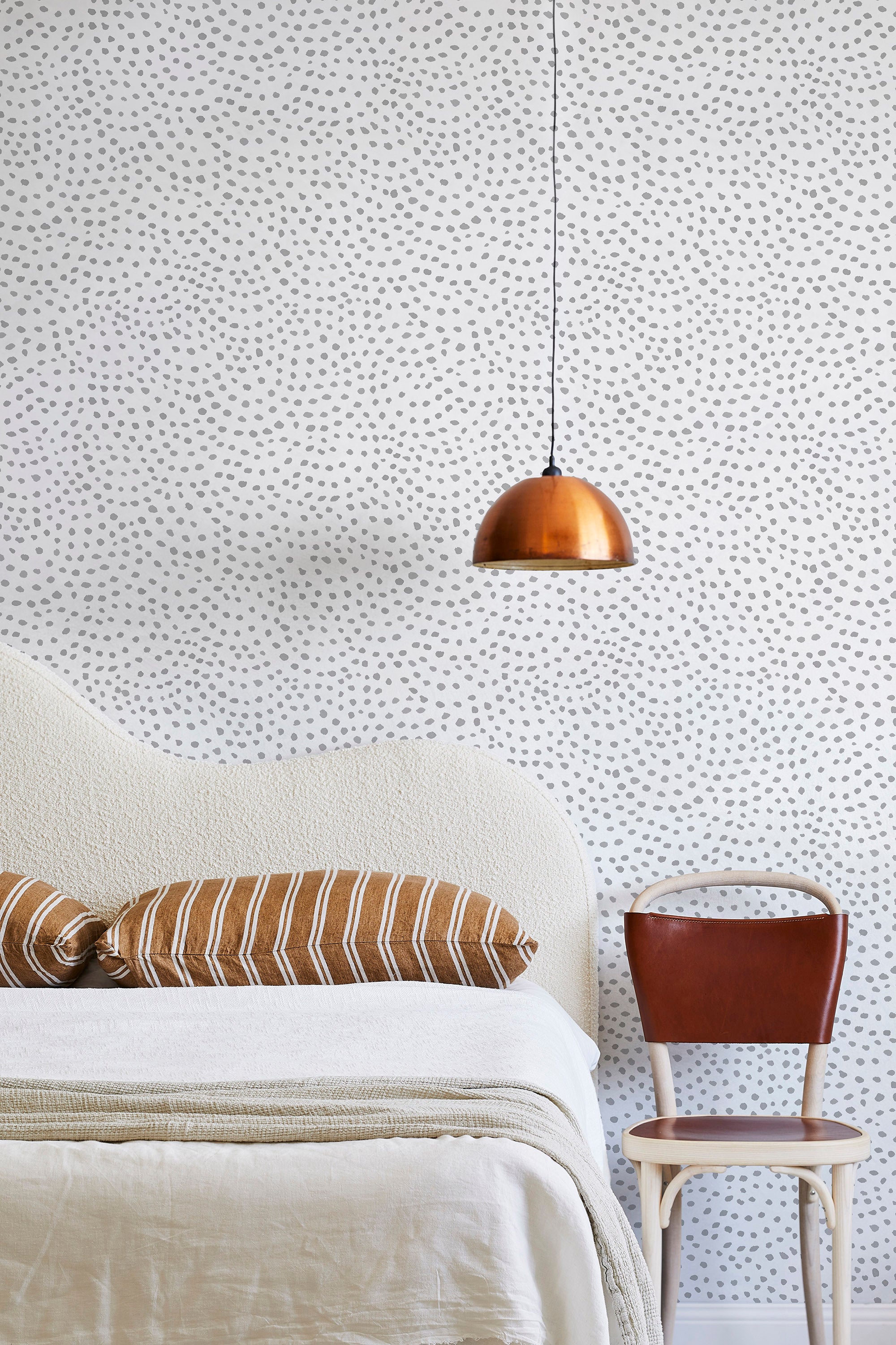 A modernist bed, hanging lamp and chair stand in front of a wall papered in a scalloped dot print in gray and cream.