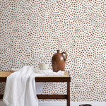 A bench with a towel and clay vases stands in front of a wall papered in a scalloped dot print in rust and cream.