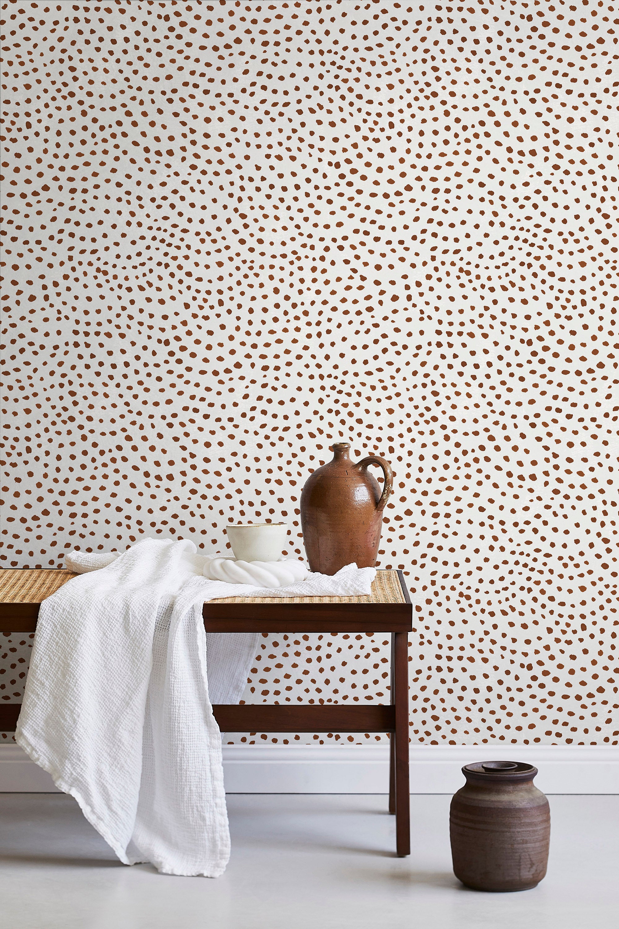 A bench with a towel and clay vases stands in front of a wall papered in a scalloped dot print in rust and cream.