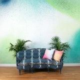 A sofa and plants stands in front of a wall covered in an abstract ombré paint splatter print in green, turquoise and white.