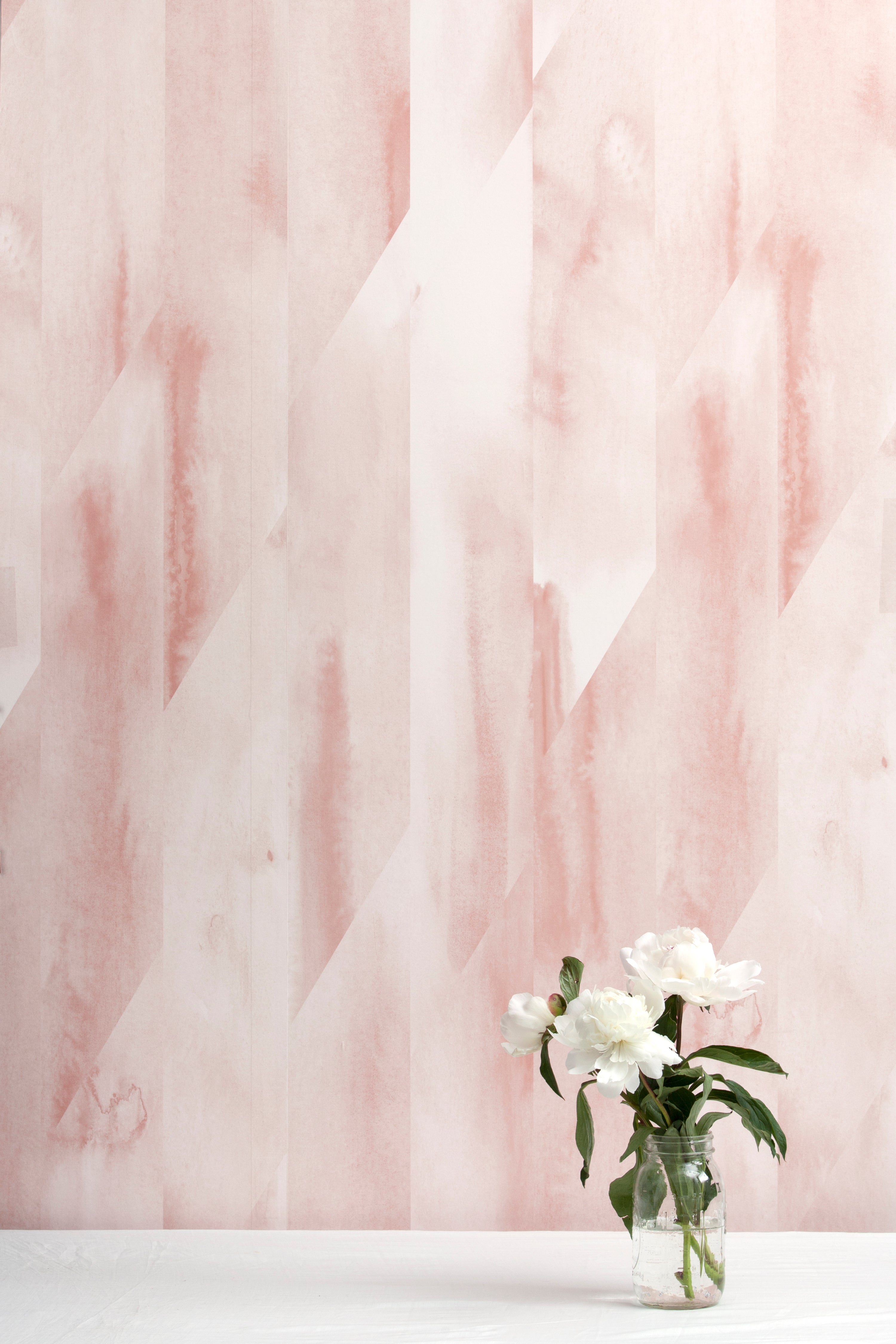 A vase of flowers stands in front of a wall papered in a linear geometric print in shades of pink and cream.