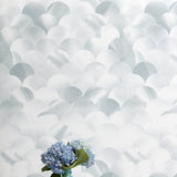 A vase of flowers stands in front of a wall papered in a repeating scalloped print in shades of gray and white.