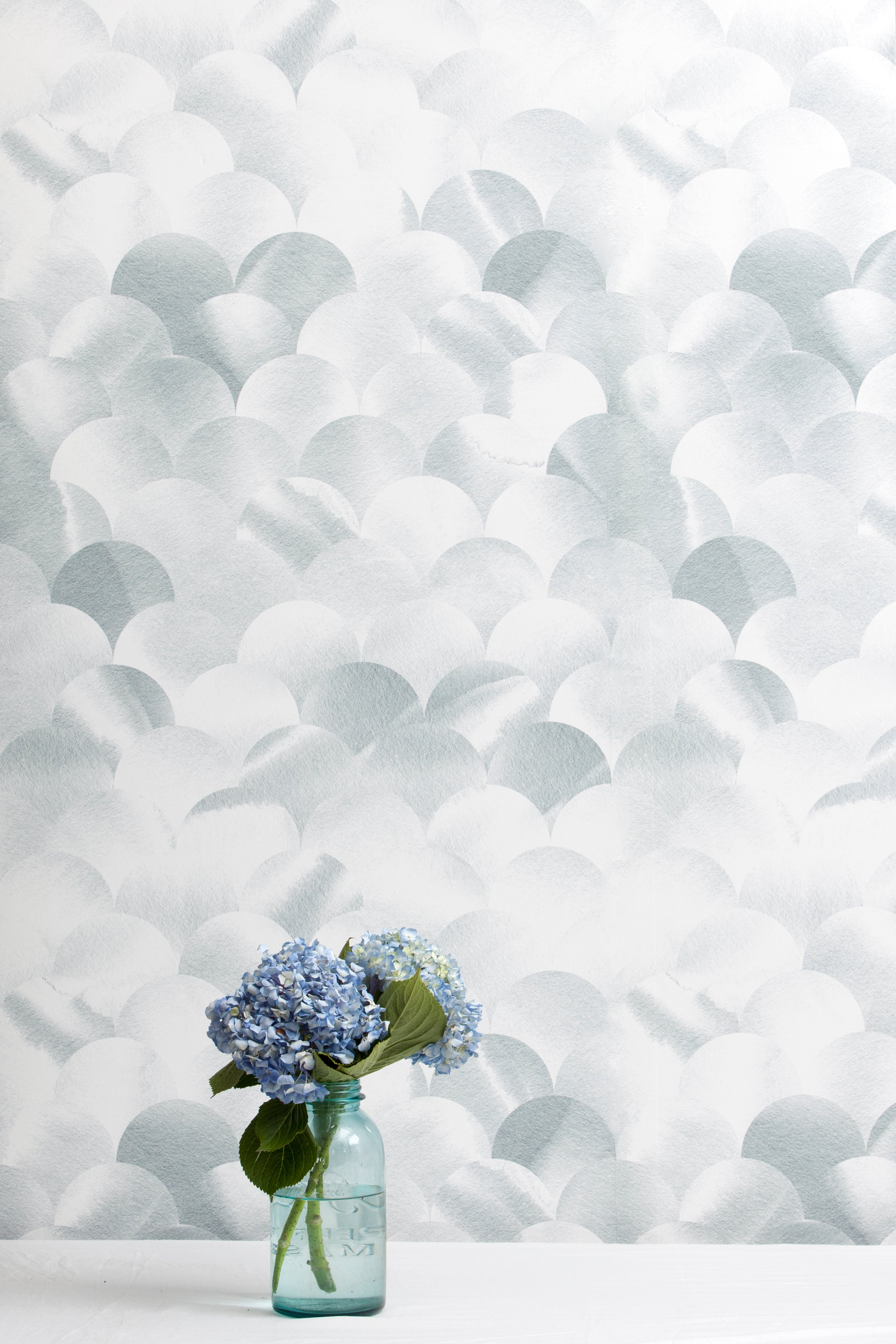 A vase of flowers stands in front of a wall papered in a repeating scalloped print in shades of gray and white.