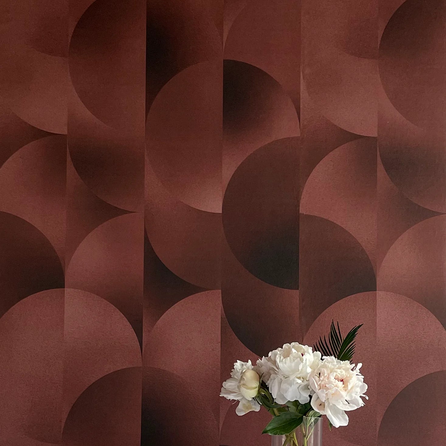 A vase of flowers stands in front of a wall covered in an abstract curvilinear print in mottled brown and red.