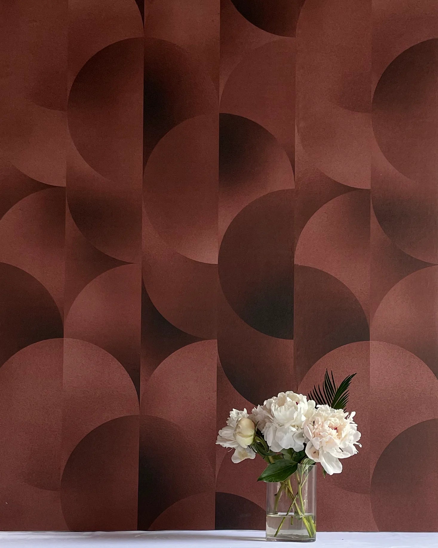 A vase of flowers stands in front of a wall covered in an abstract curvilinear print in mottled brown and red.