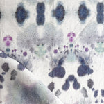 Draped fabric yardage in an abstract inkblot print in shades of purple and gray on a cream field.