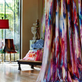 A long curtain in an abstract painterly print in shades of pink, purple and blue hangs in front of a cluttered living space.