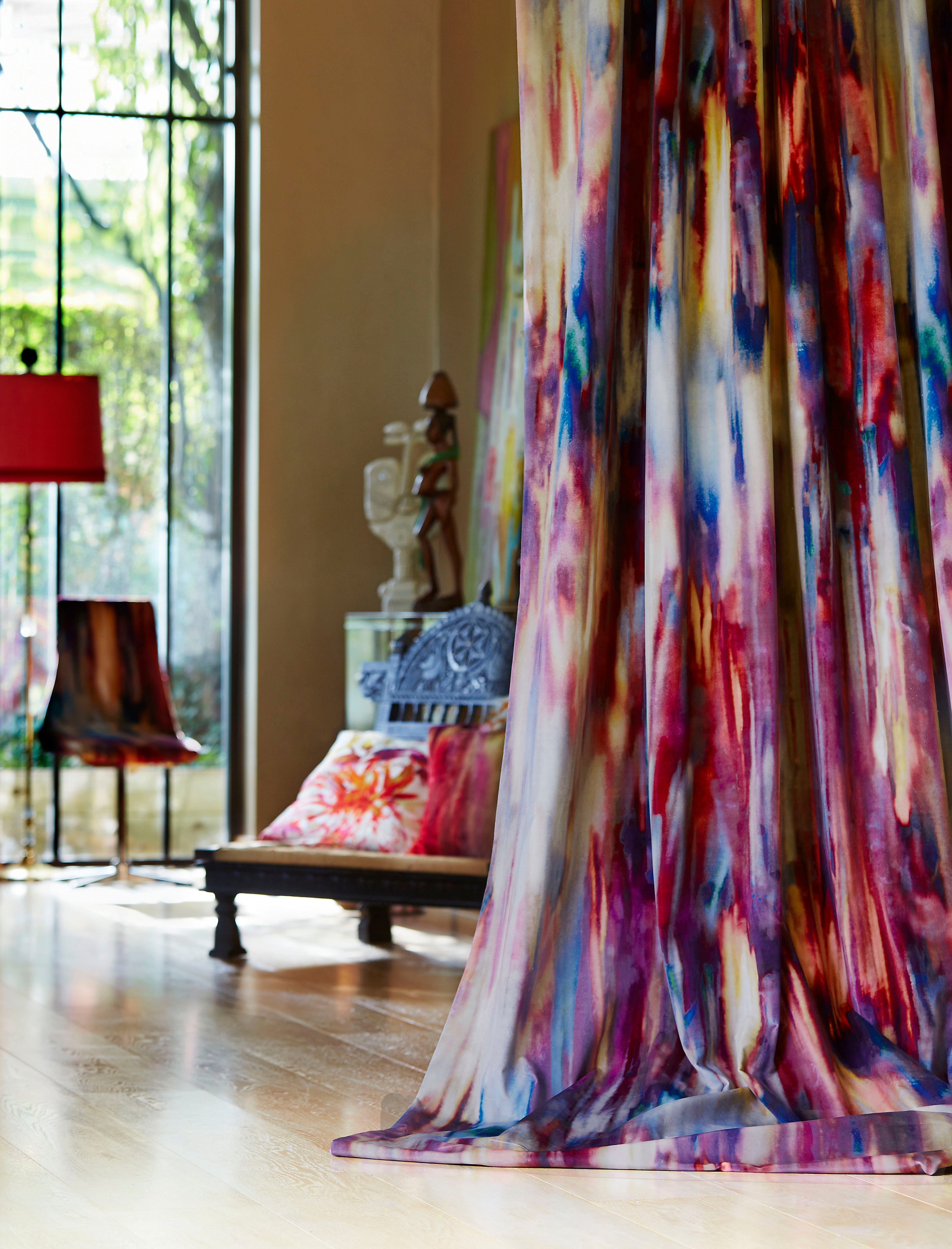 A long curtain in an abstract painterly print in shades of pink, purple and blue hangs in front of a cluttered living space.