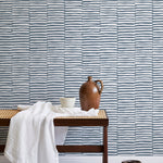 A bench with a towel and clay vases stands in front of a wall papered in a broken stripe print in navy and white.