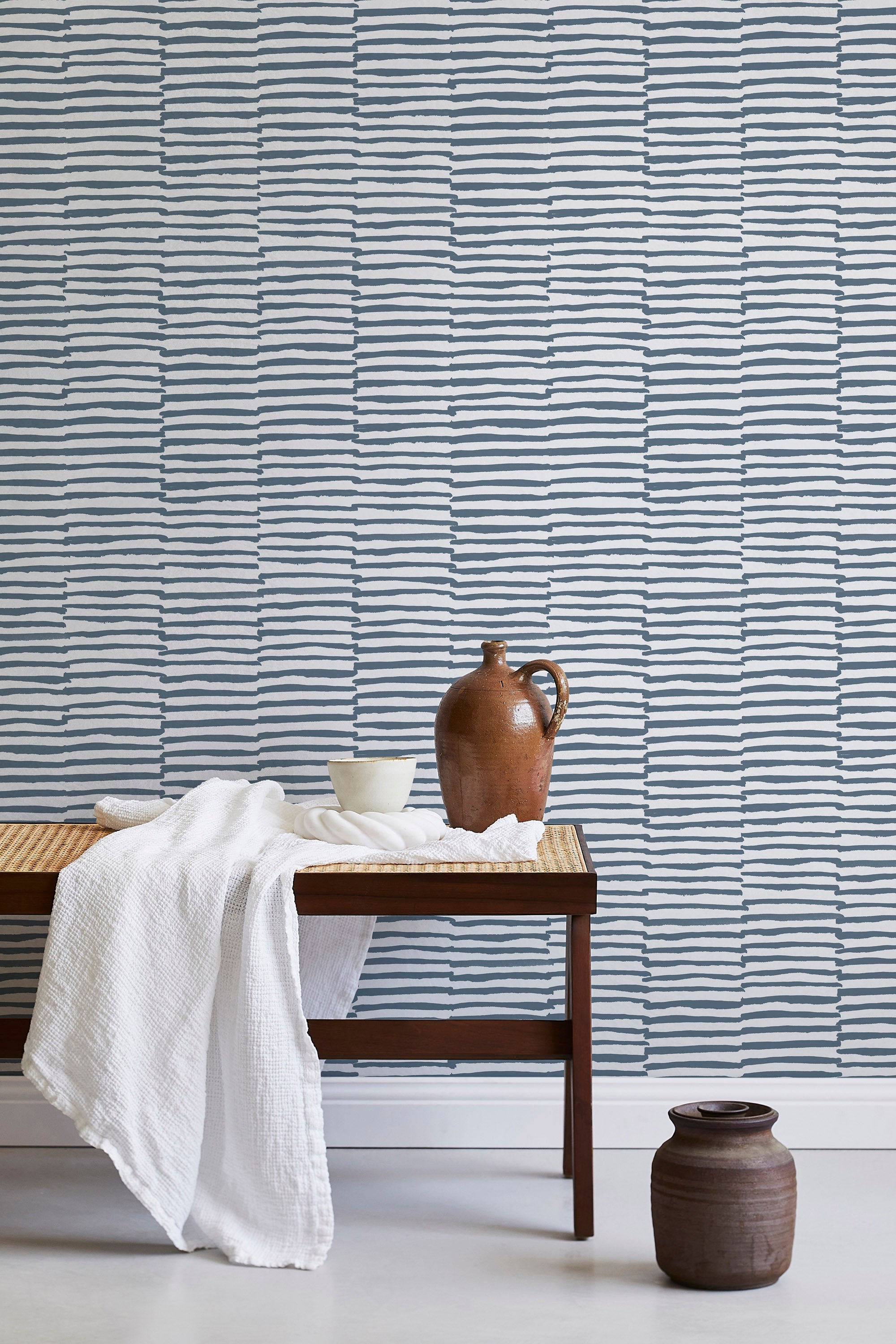 A bench with a towel and clay vases stands in front of a wall papered in a broken stripe print in navy and white.