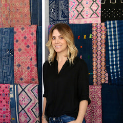 A woman with blonde hair wearing jeans and a black top stands in front of a background of patchworked interior fabrics.