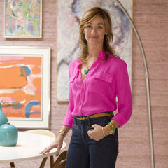 A blonde woman in a pink button-down, layered gold jewelry and jeans stands in front of a mauve wall covered in paintings.
