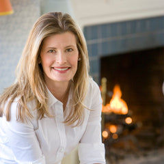 Close-up of a blonde woman wearing a white buttondown and smiling, seated in front of a fireplace.