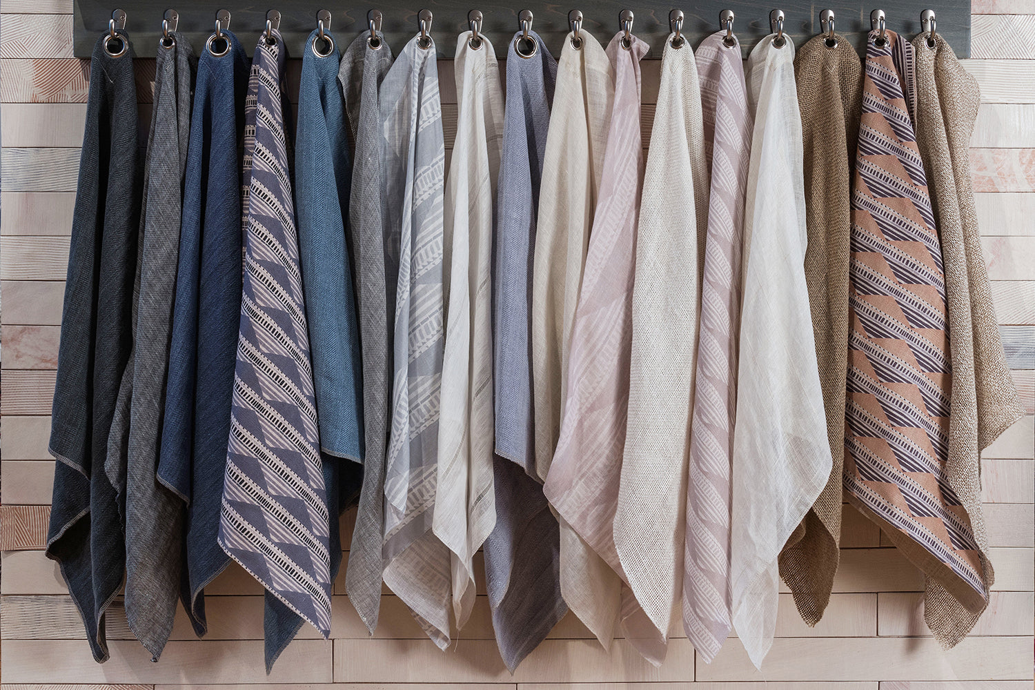 A row of fabric cuttings, hung on on a rack with hooks in a range of charcoal, navy, slate, grey, ivory, beige and tan. 