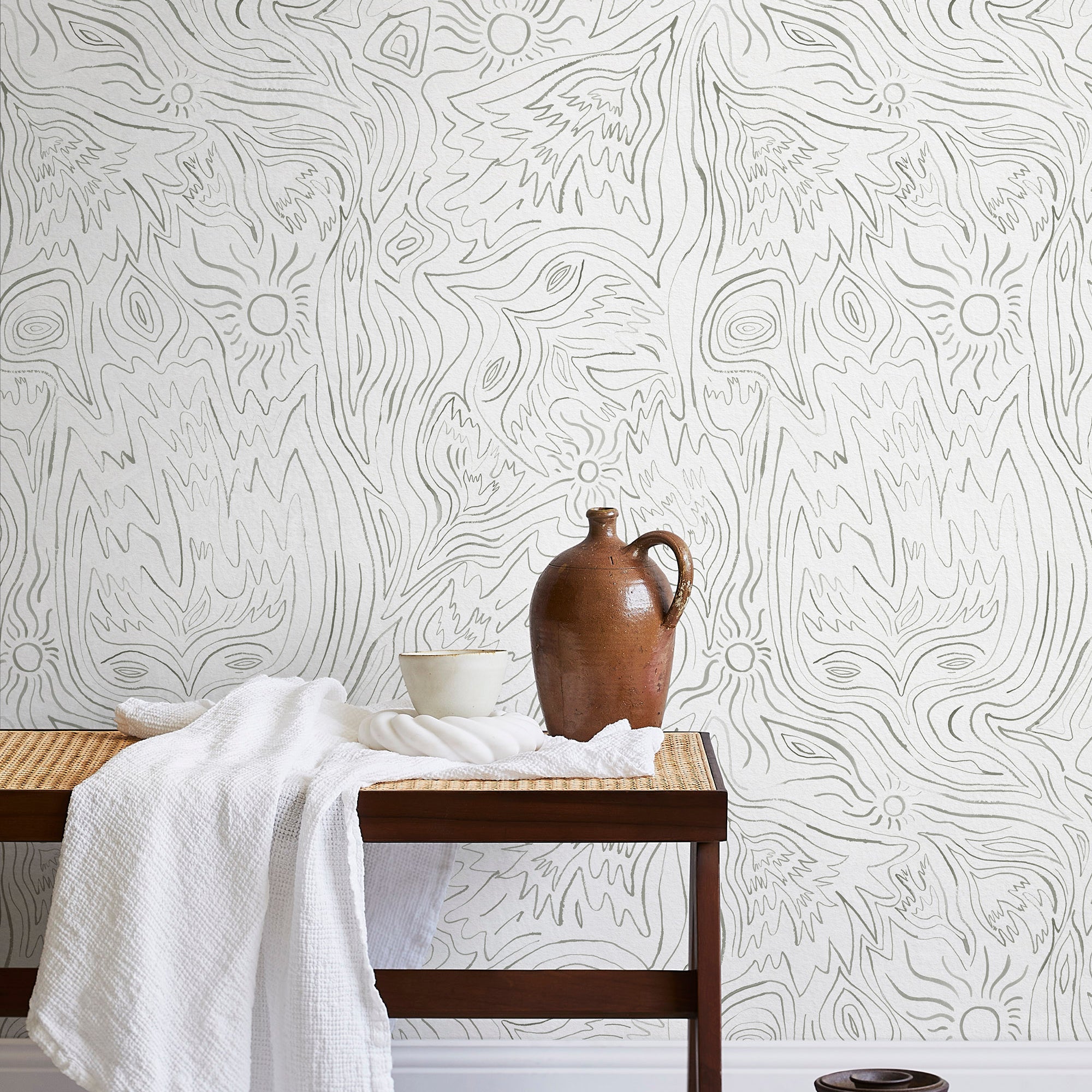 A bench with a towel and clay vases stands in front of a wall papered in a playful abstract sun print in gray and cream.