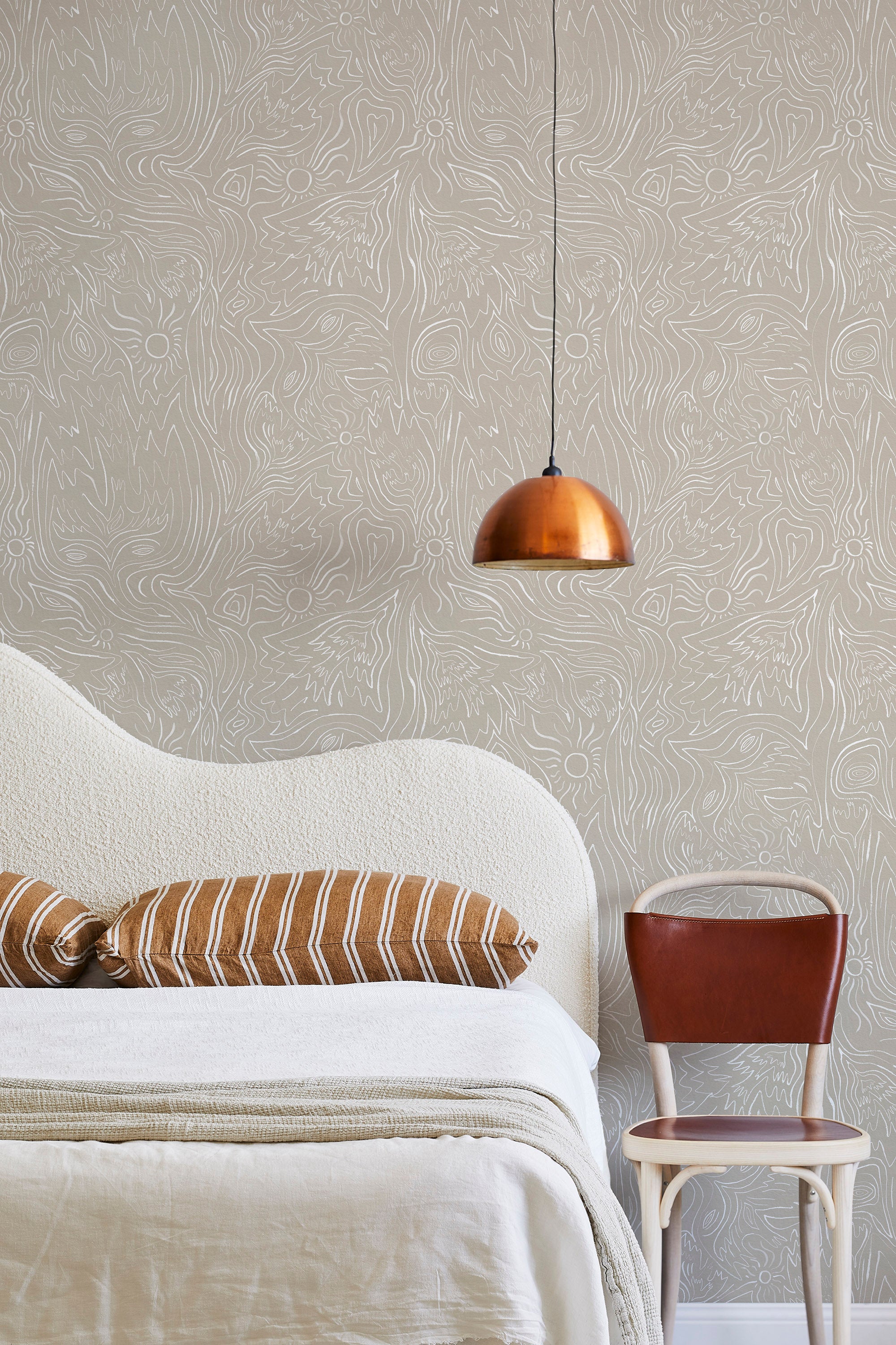 A modernist bed, hanging lamp and chair stand in front of a wall papered in a playful abstract sun print in white and cream.