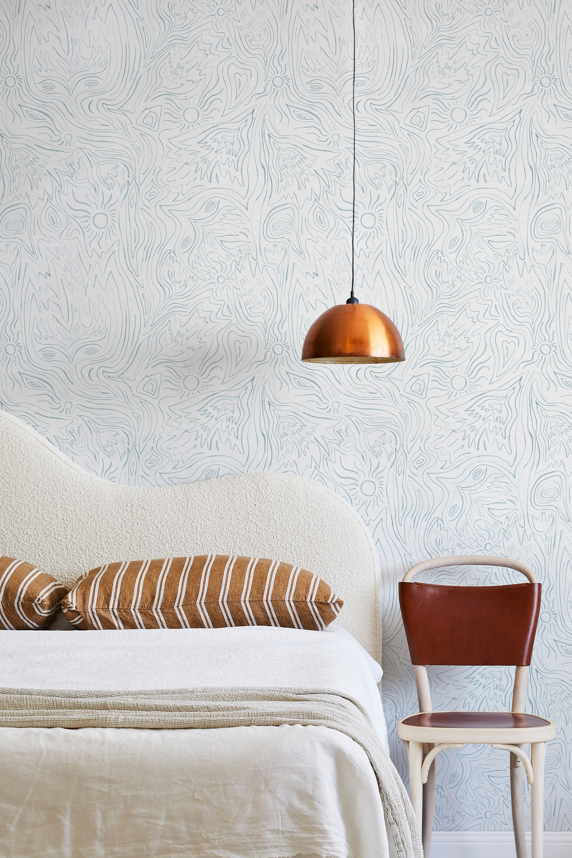 A modernist bed, hanging lamp and chair stand in front of a wall papered in an abstract sun print in light blue and white.