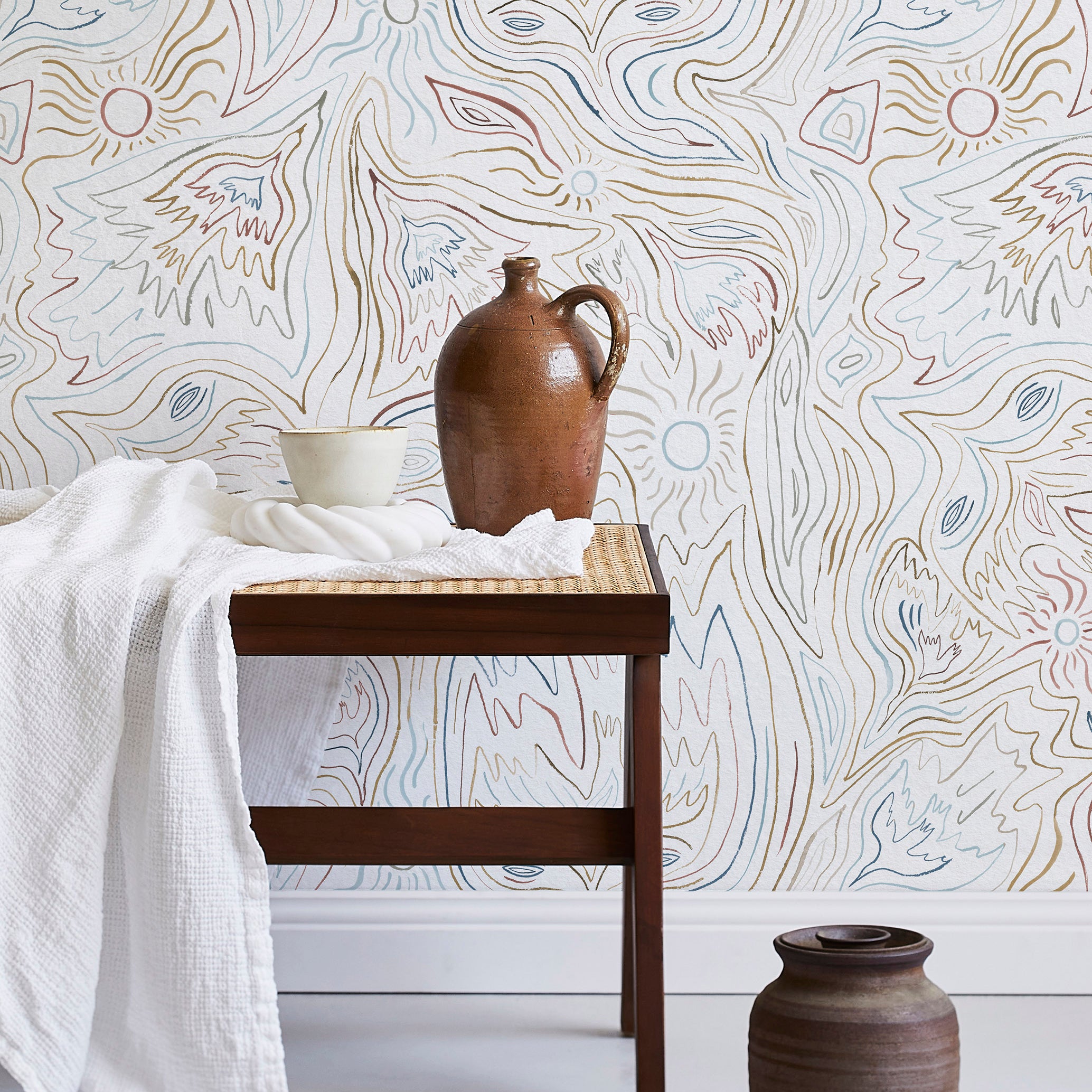 A bench with a towel and clay vases stands in front of a wall papered in an abstract sun print in brown, blue and white.