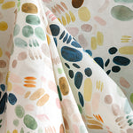 Draped fabric yardage in a playful paint blotch print in shades of blue, yellow and pink on a cream field.