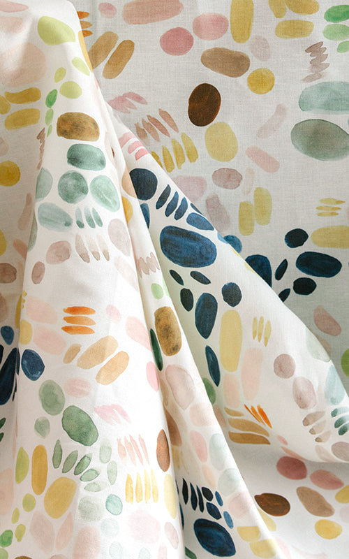 Draped fabric yardage in a playful paint blotch print in shades of blue, yellow and pink on a cream field.