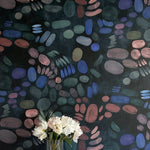A vase of flowers stands in front of a wall covered in a playful paint blotch print in blue, green and pink on a black field.