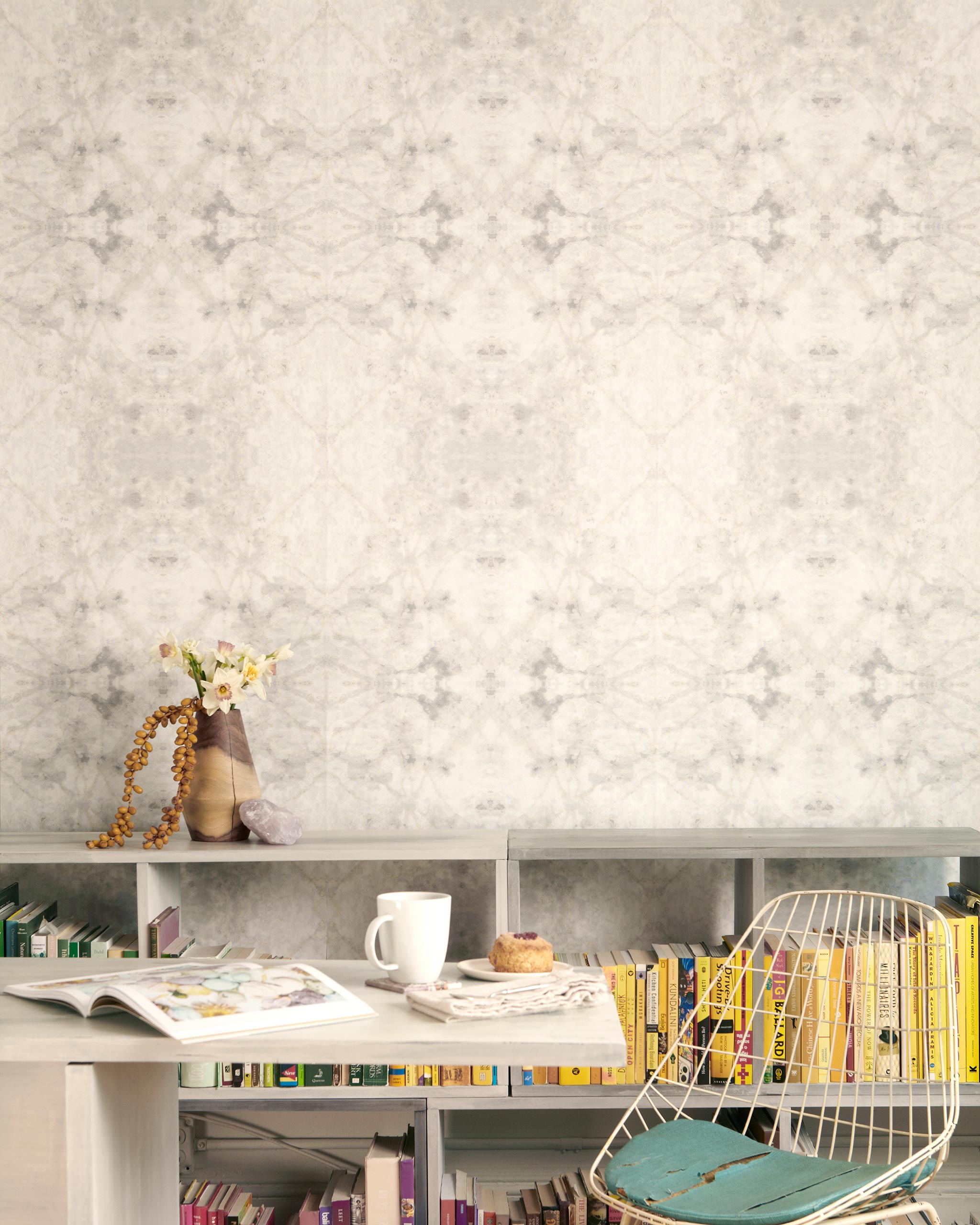 A cluttered desk and bookshelf stand in front of a wall papered in an abstract textural print in cream, gray and tan.