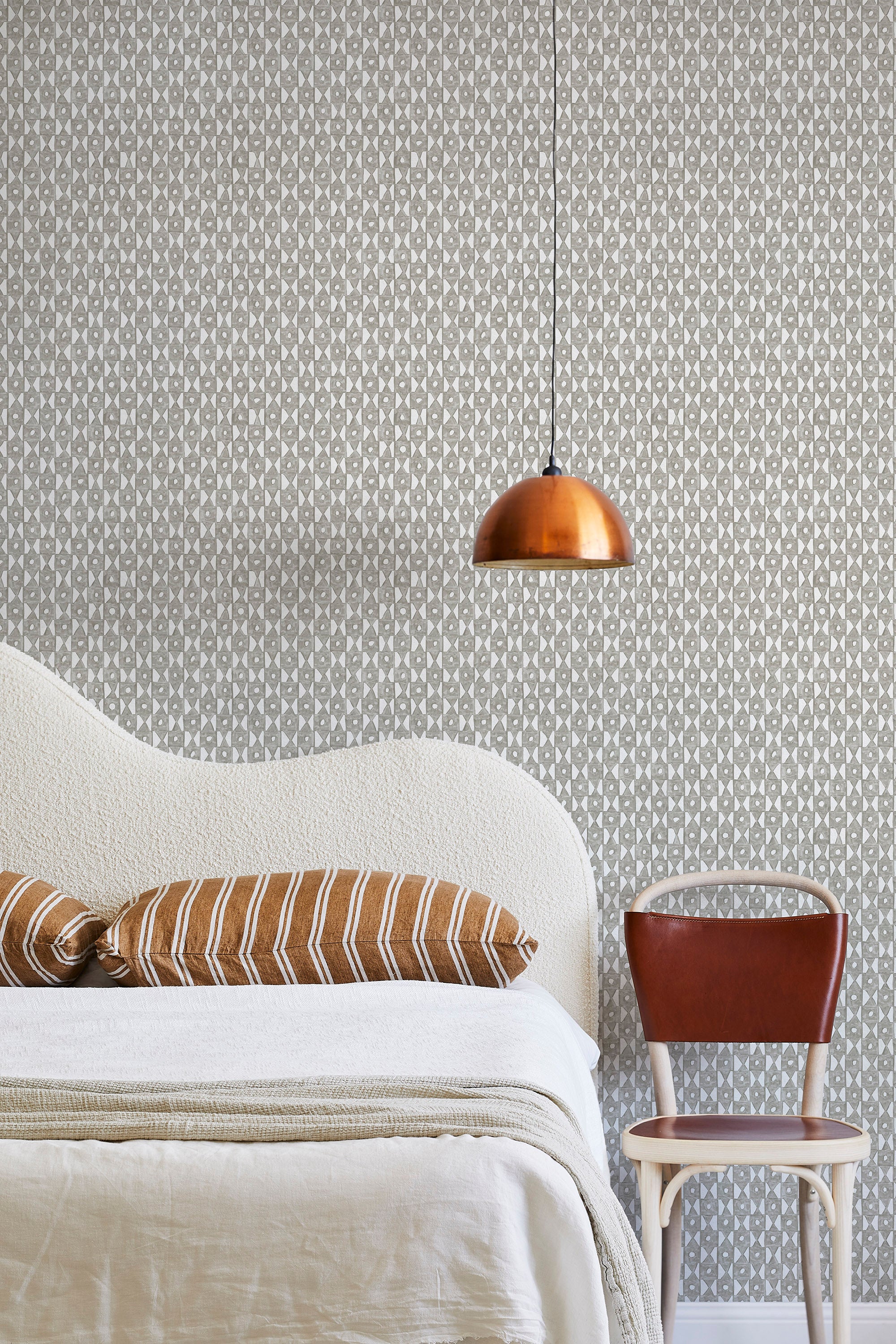 A modernist bed, hanging lamp and chair stand in front of a wall papered in a geometric grid print in gray and white.