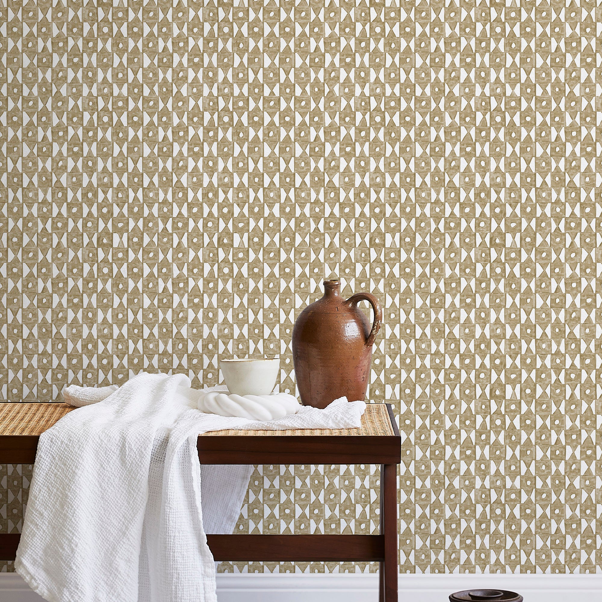 A bench with a towel and clay vases stands in front of a wall papered in a geometric grid print in brown and white.