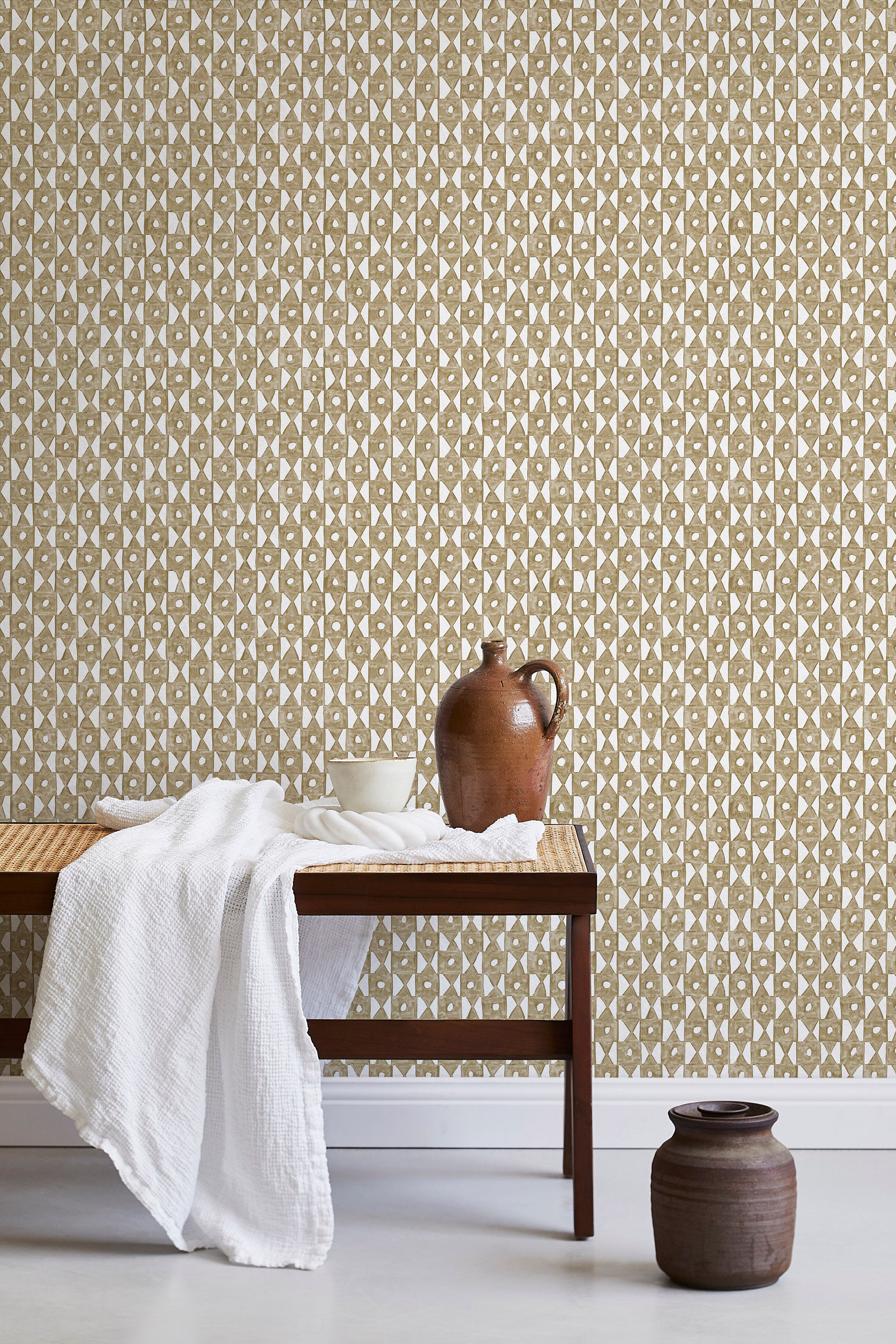 A bench with a towel and clay vases stands in front of a wall papered in a geometric grid print in brown and white.