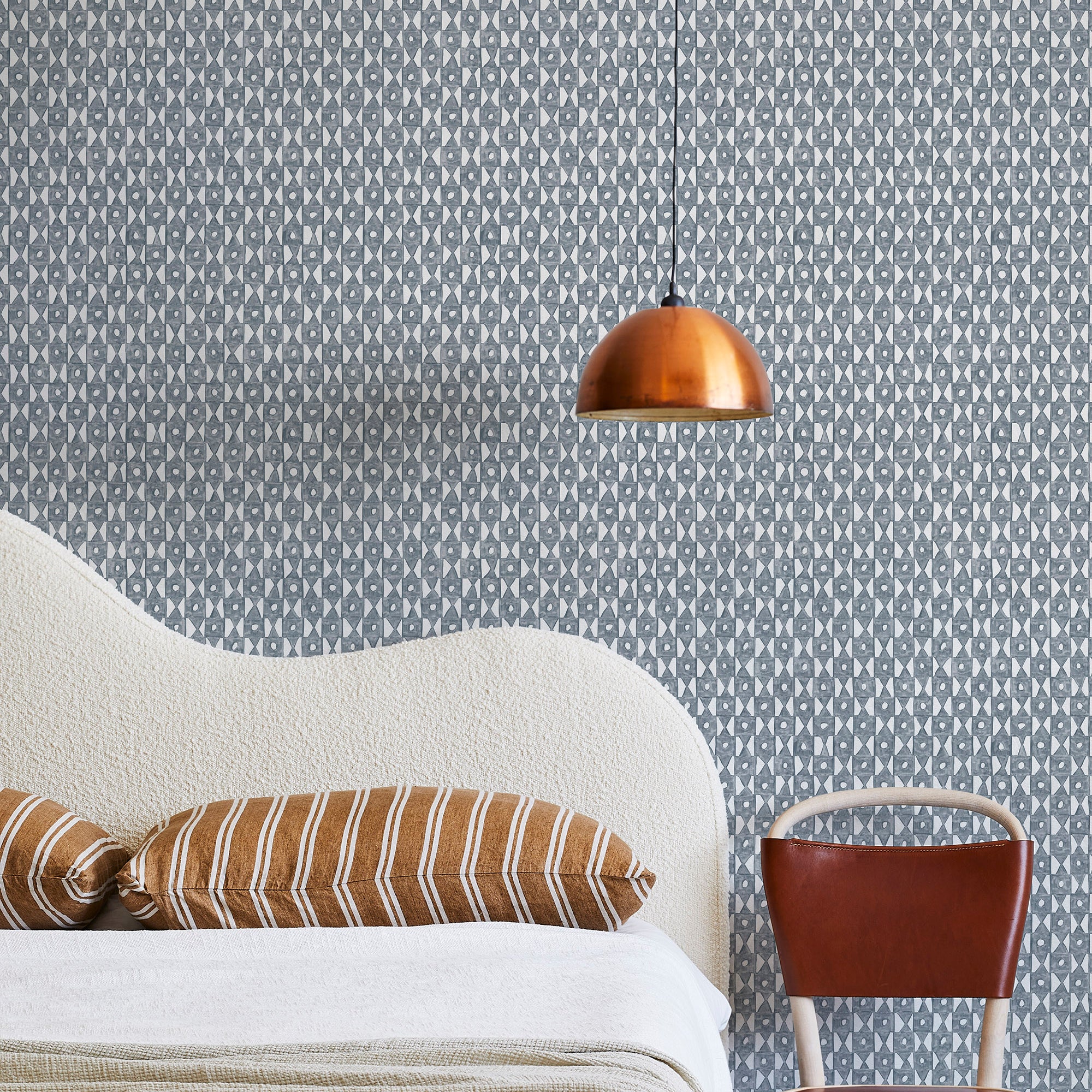 A modernist bed, hanging lamp and chair stand in front of a wall papered in a geometric grid print in navy and white.