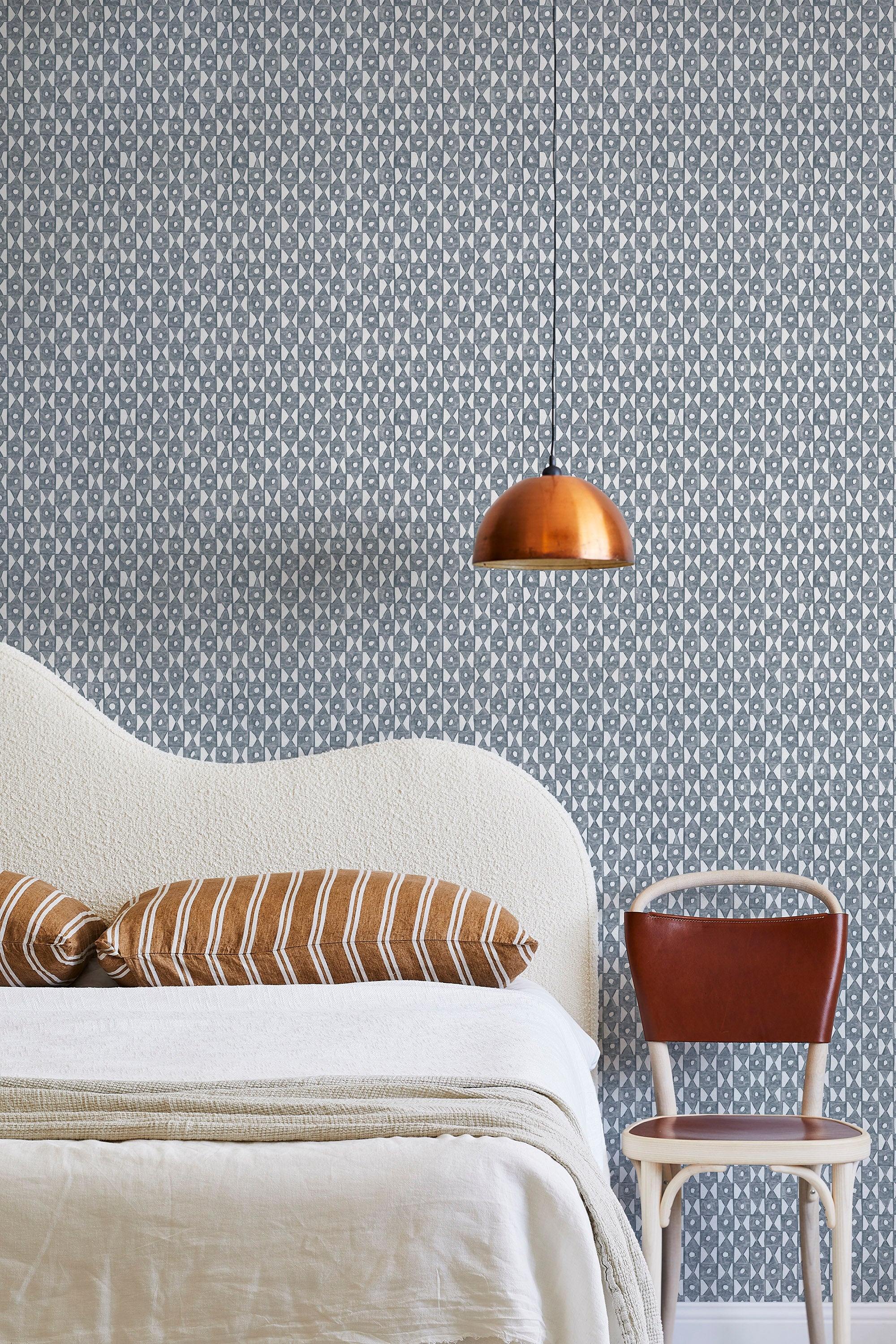 A modernist bed, hanging lamp and chair stand in front of a wall papered in a geometric grid print in navy and white.