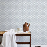 A bench with a towel and clay vases stands in front of a wall papered in a geometric grid print in light blue and white.