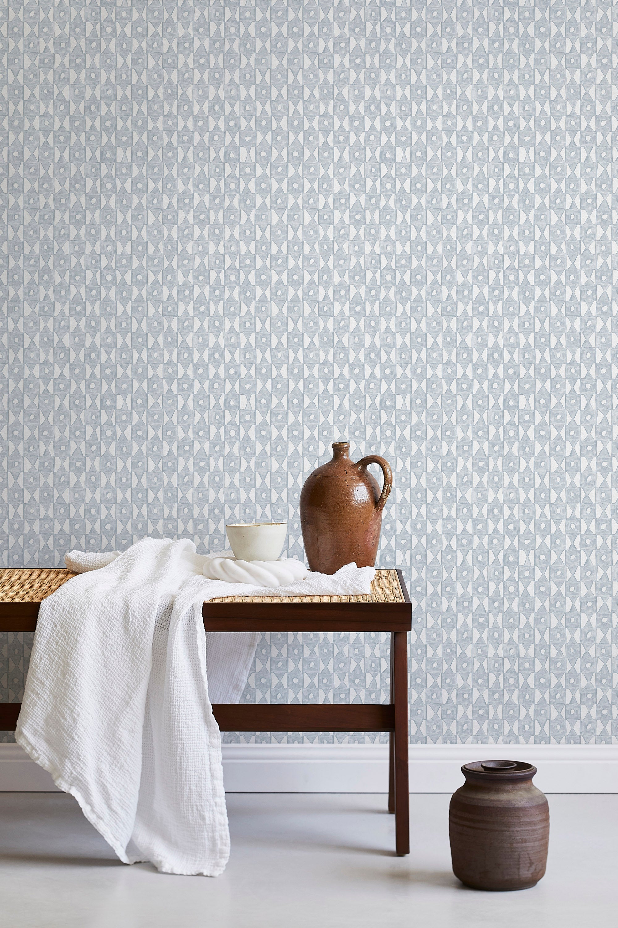 A bench with a towel and clay vases stands in front of a wall papered in a geometric grid print in light blue and white.