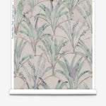 Partially unrolled wallpaper yardage in a painterly palm tree pattern in shades of green and purple on a tan field.